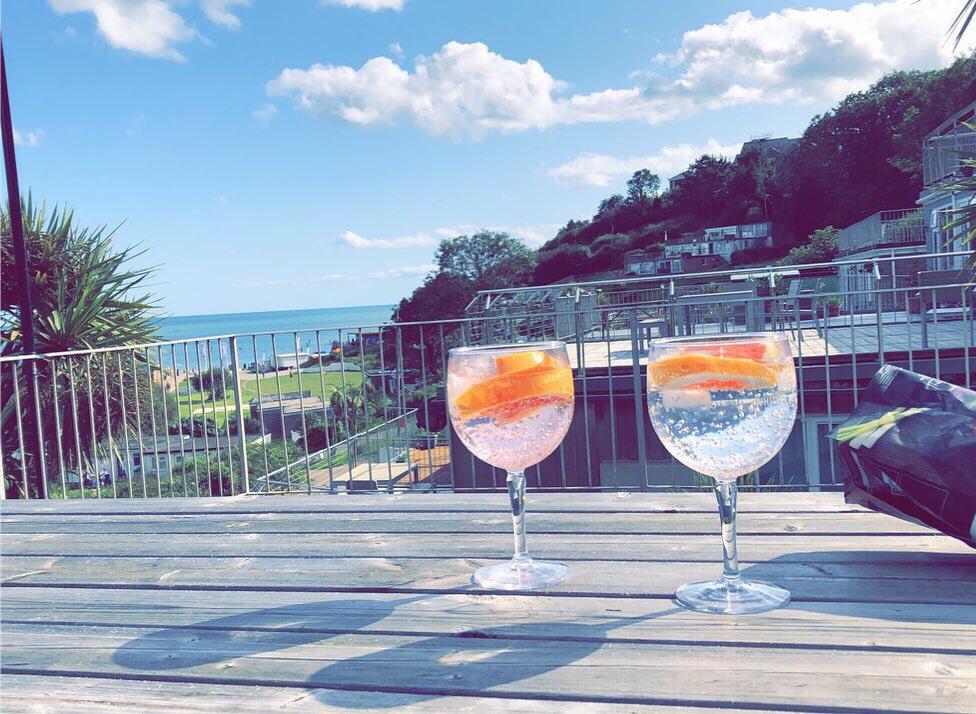 Two glasses of sparkling water with orange slices are positioned on a wooden table. A coastal view is visible in the background, with lush greenery and blue waters under a partly cloudy sky.