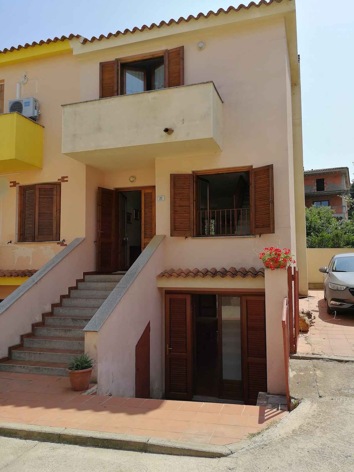 The entrance to the building is framed by a staircase leading to the main door, flanked by wooden shutters. A small balcony is visible above, with flowering plants adorning the entrance. The exterior portrays a pastel color, complemented by a parking area for vehicles.