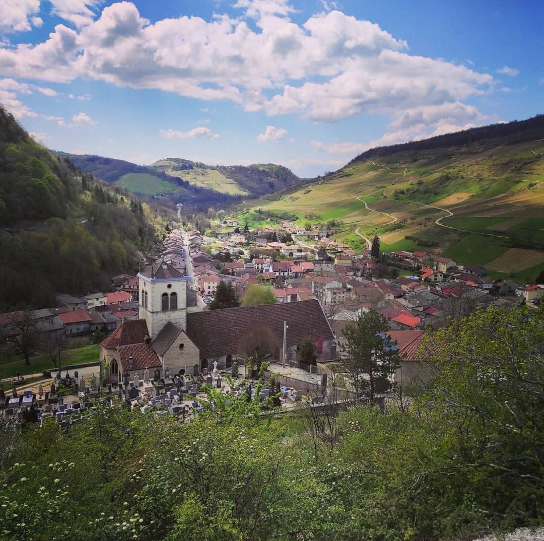 A panoramic view of a small village nestled in a valley, with green hills rising in the background. The image highlights a church with a bell tower in the foreground, surrounded by traditional buildings and a cemetery, showcasing the tranquil rural landscape.