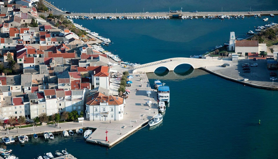 An aerial view captures the marina, showcasing several docked boats along the turquoise water. The waterfront is lined with buildings featuring red-tiled roofs, while a distinctive white bridge arches over the water, leading to a small harbor area.