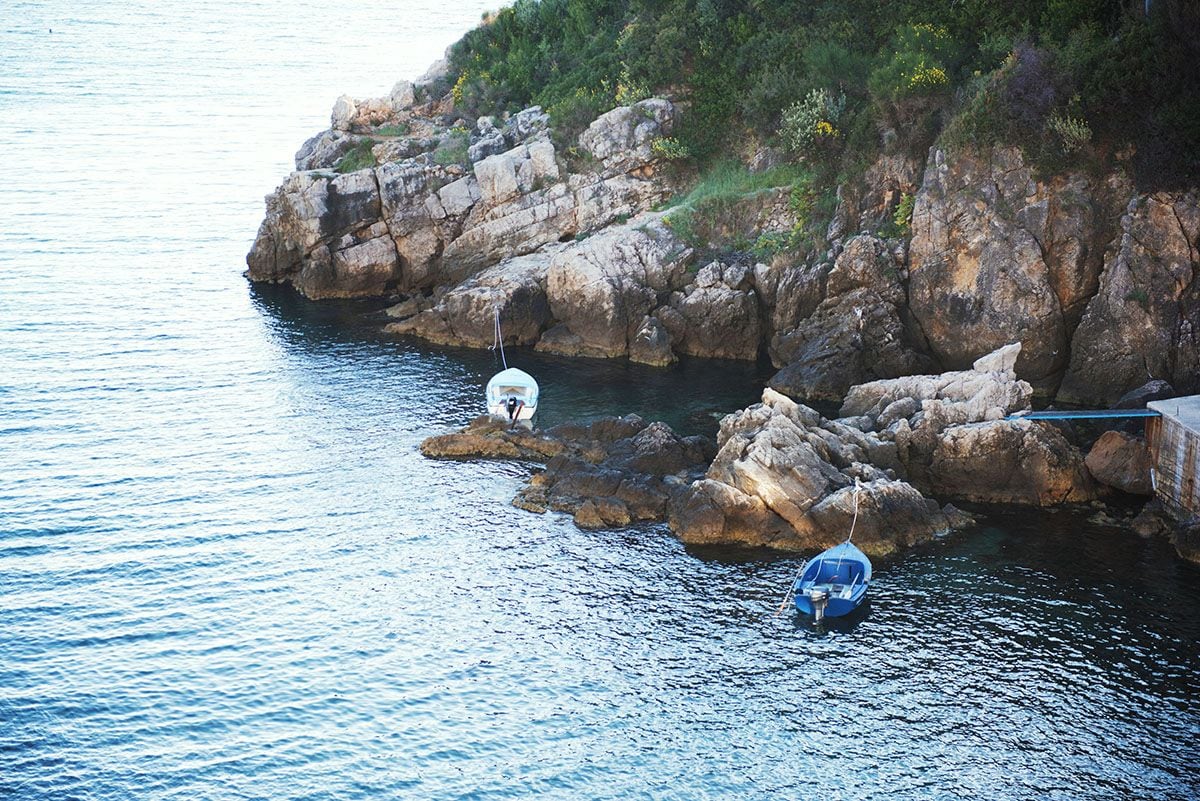 Two boats are moored near rocky shoreline, surrounded by calm water. The natural landscape features rugged cliffs, with greenery visible on the hillside, creating a serene coastal setting.