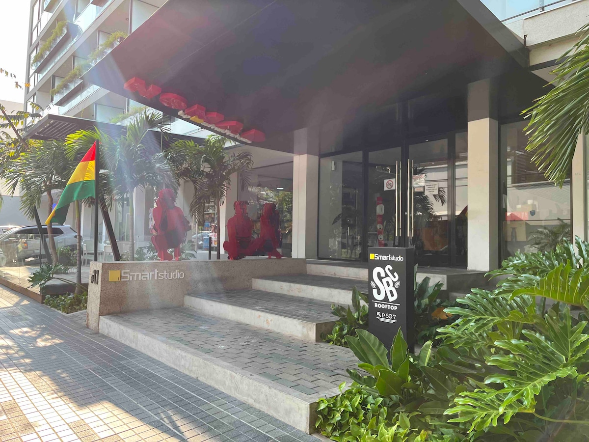 A modern building entrance is framed by lush greenery, featuring steps leading up to a spacious lobby with large glass doors. The property's signage is visible, complemented by decorative plants and a flagpole displaying the national flag.