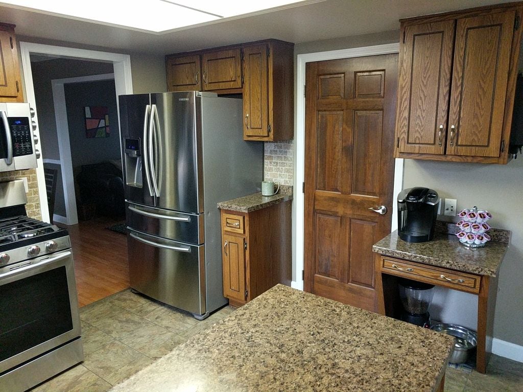 The kitchen features wooden cabinetry and a large granite countertop. Stainless steel appliances include a refrigerator and a gas range. A Keurig coffee maker is positioned on a smaller counter, while a doorway leads to a living area, creating an open and functional layout.