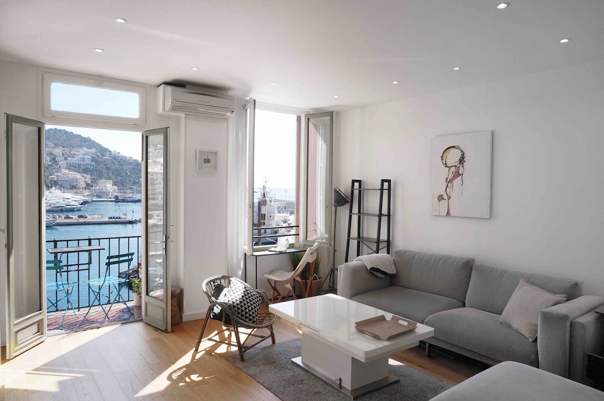 A bright living area features large windows that open to a balcony, showcasing views of the Mediterranean Sea. Comfortable seating includes a spacious couch and a stylish chair. Air conditioning and modern décor are present, along with a small white coffee table and neutral-toned rug.