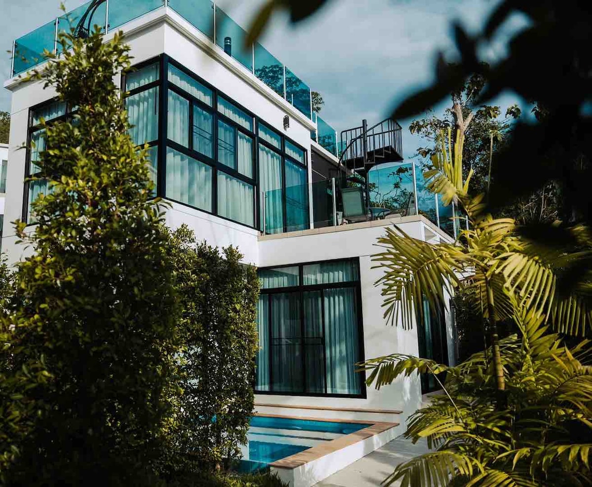 The villa's modern design is showcased through expansive glass windows that reflect the surrounding greenery. A private plunge pool is visible in the foreground, with tropical plants framing the property. The rooftop terrace offers additional outdoor space, enhancing the villa's appeal.