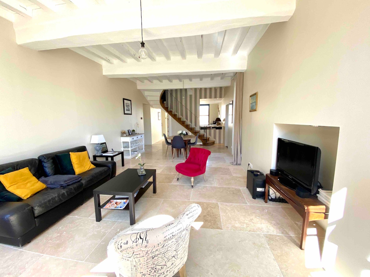 A spacious living area features a dark sectional sofa with yellow cushions, accent chairs, and a low coffee table. Light from large windows illuminates the room, highlighting the stone flooring and a wooden staircase leading to the upper level.
