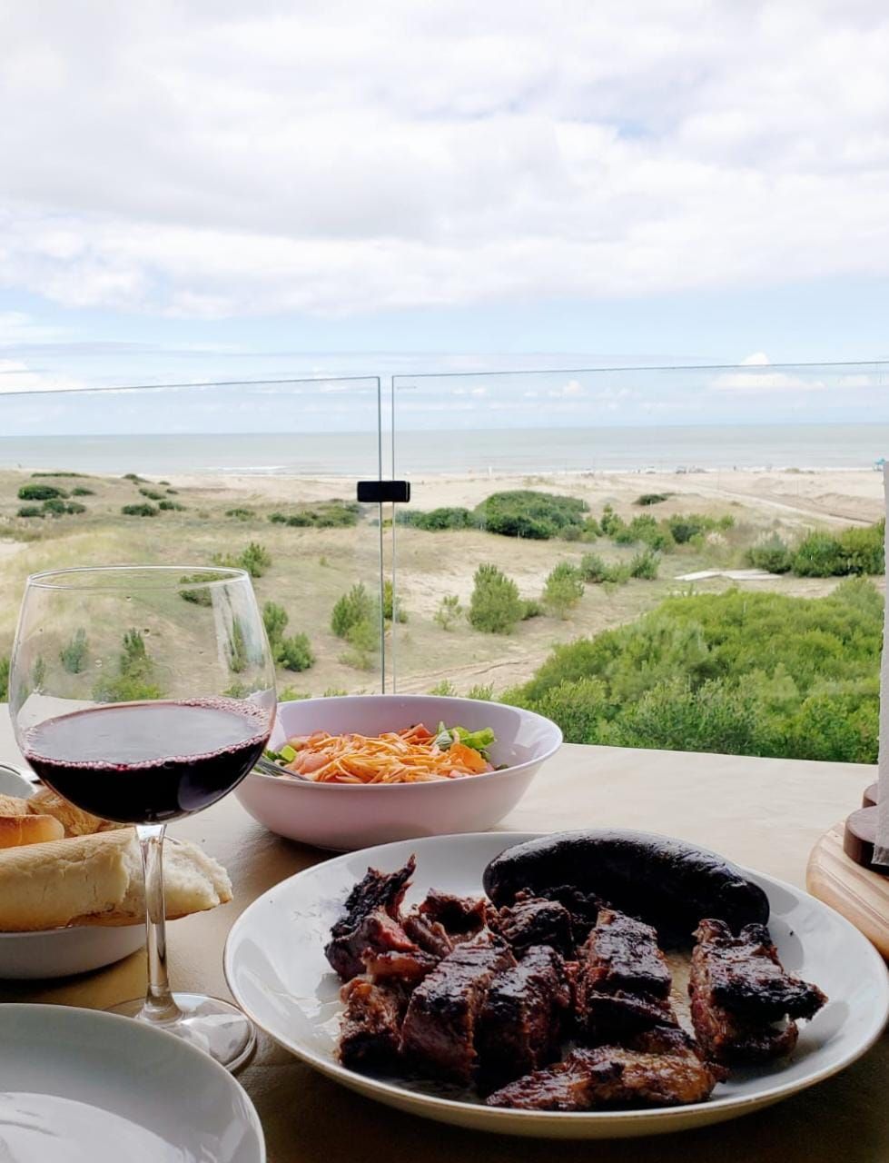 A dining table is set with an array of dishes, including grilled meat and fresh salads. A glass of red wine sits beside the plates. The backdrop features a scenic view of the ocean and greenery through a glass railing.