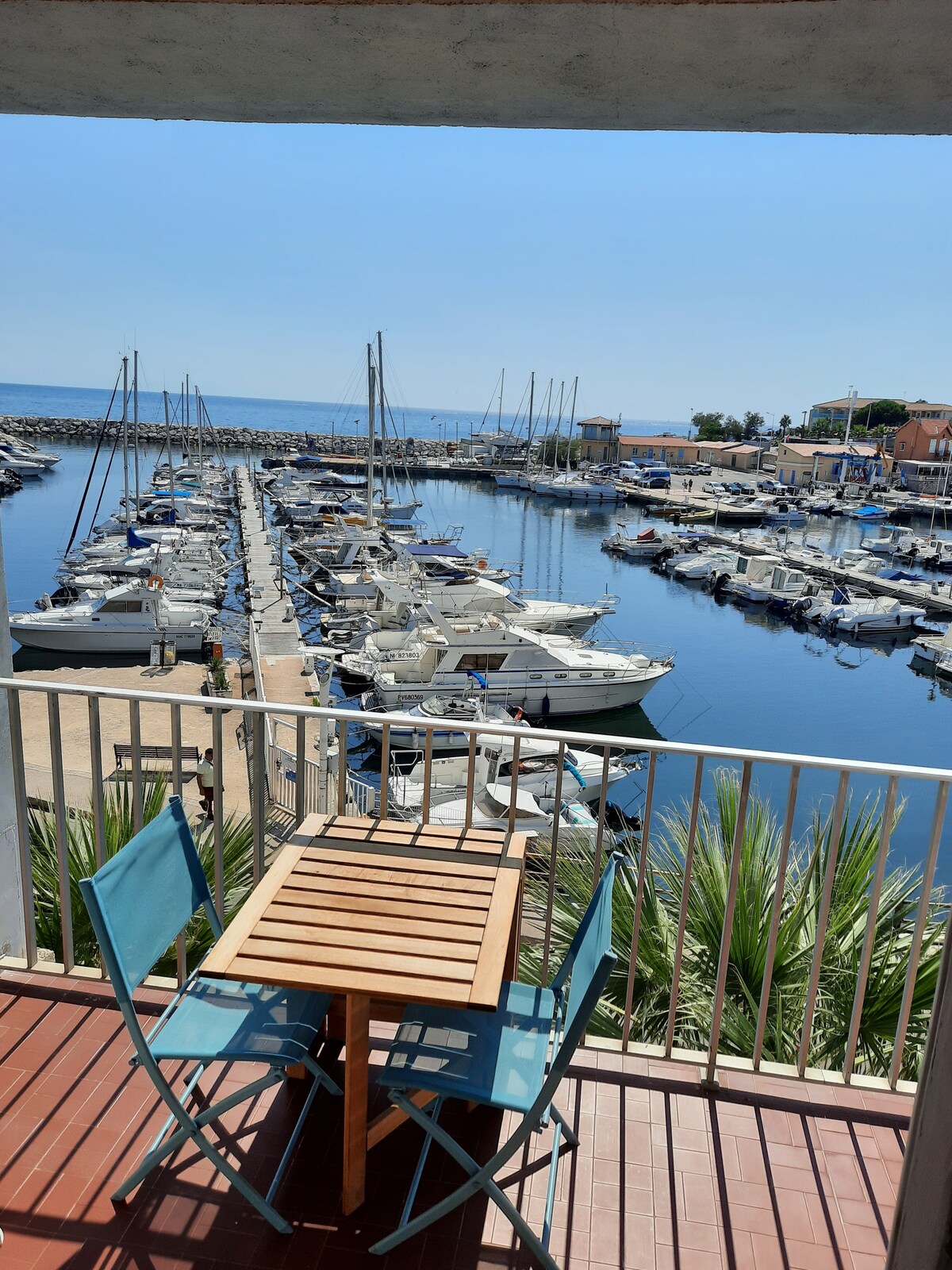 A view from the terrace showcases the marina filled with boats nestled along the calm waters. Small blue chairs and a wooden table are positioned on the terrace, providing a cozy space to enjoy the scenery under the clear sky.