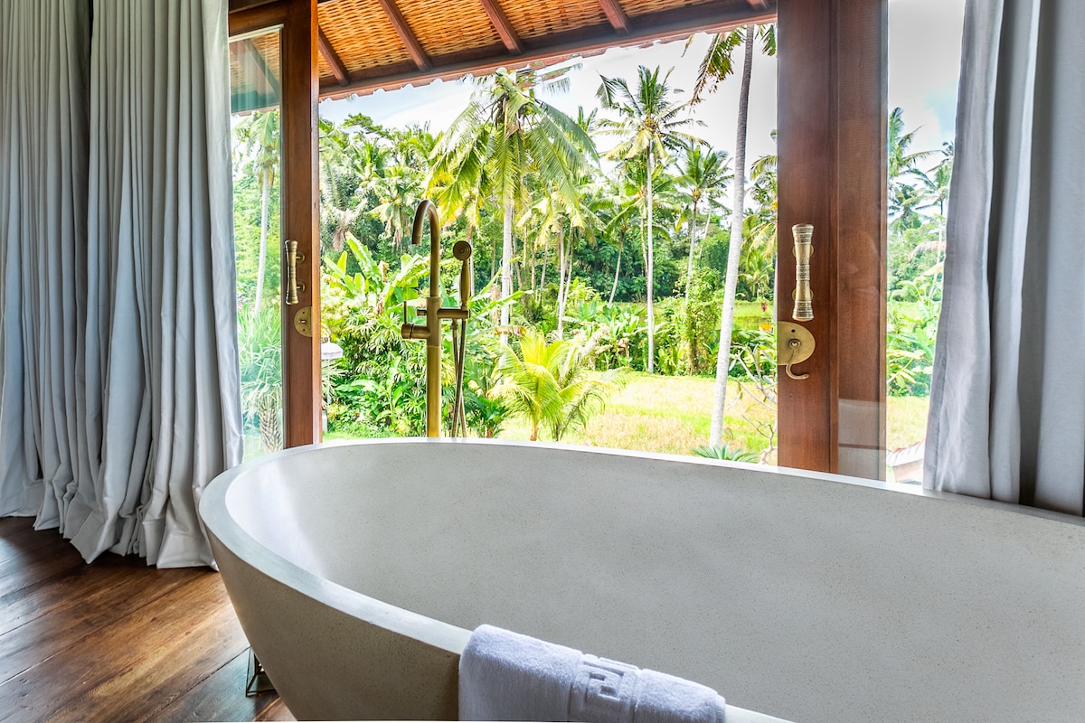 A freestanding bathtub is positioned by expansive windows, offering views of lush greenery. The natural light enhances the serene atmosphere, while soft curtains frame the scene. A rolled towel rests on the tub's edge, contributing to the inviting spa-like setting.