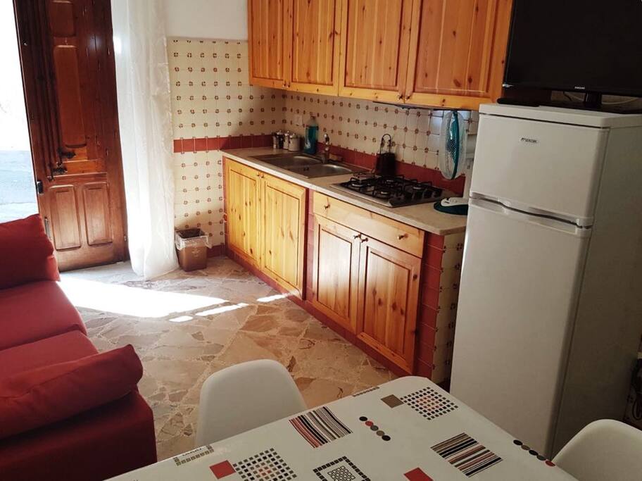 Cucina/soggiorno composta da: tavolo in legno con 4 sedie, cucina con fornelli, TV, lavandino, frigorifero e divano