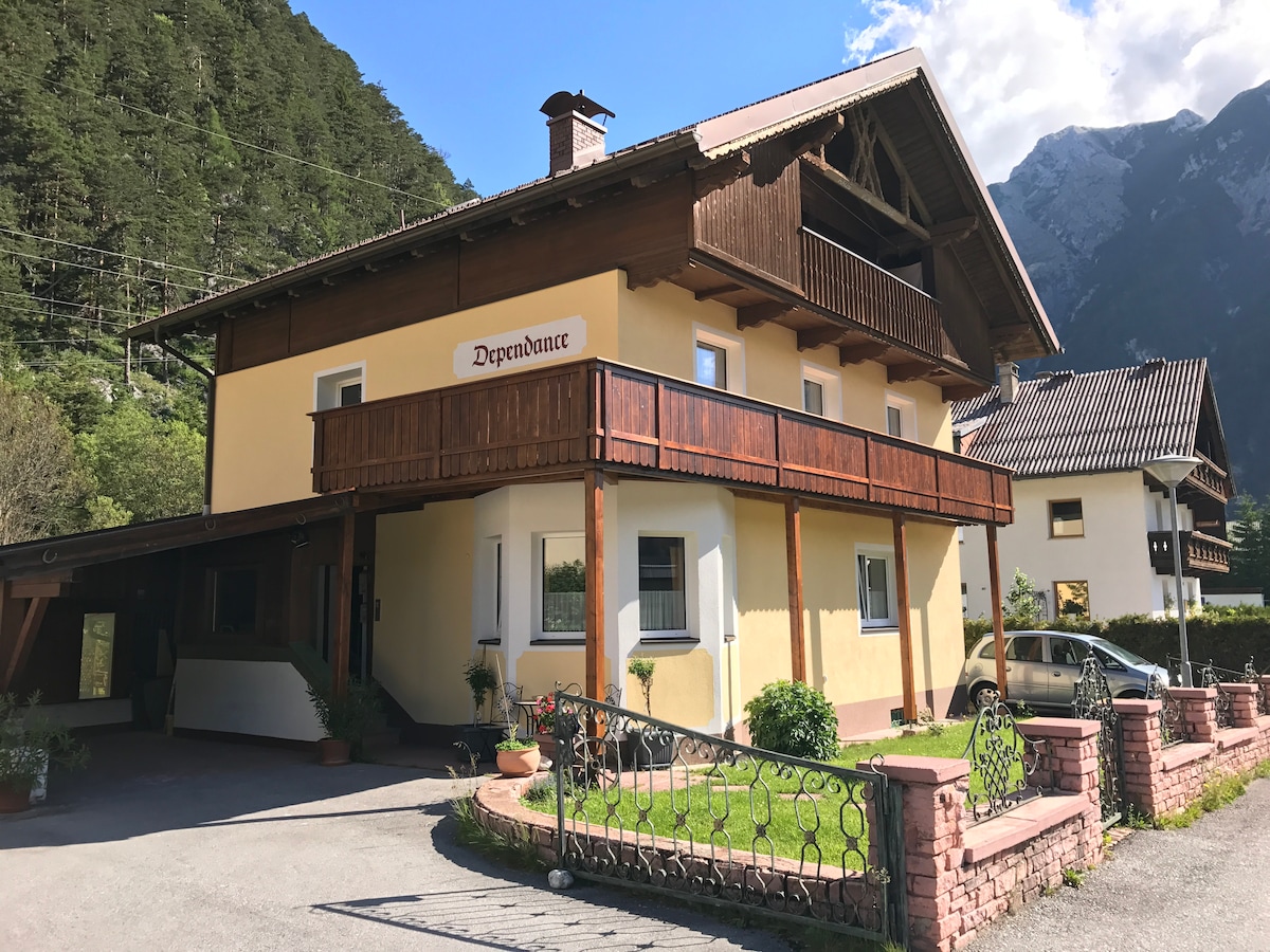 The two-story building features a warm yellow exterior with wooden accents, including a prominent balcony. A well-maintained garden with potted plants is visible in the foreground, while a driveway accommodates a parked car. Lush green hills and mountains form a scenic backdrop.