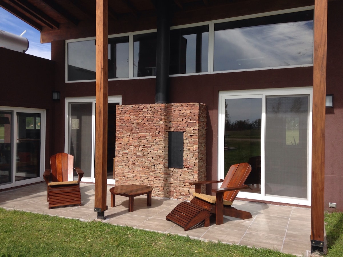 An outdoor seating area features two wooden chairs and a circular table set on tiled flooring. A stone fireplace is positioned against the wall, with large windows providing views of the surrounding landscape and natural light entering the space.