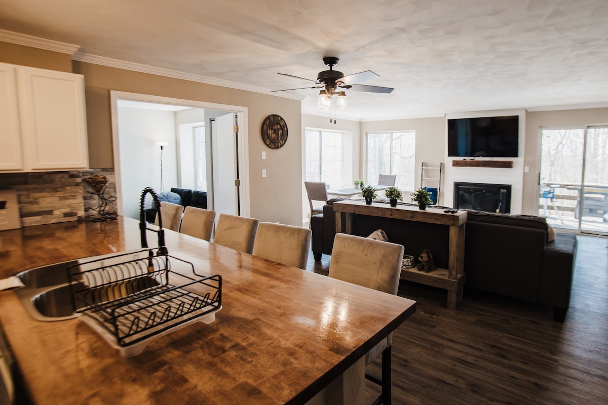 The open living space features a large dining table with light-colored chairs and an adjoining kitchen with a wooden countertop. A comfortable sectional sofa faces a TV, and large windows allow natural light to fill the room, offering a view of the outdoors.