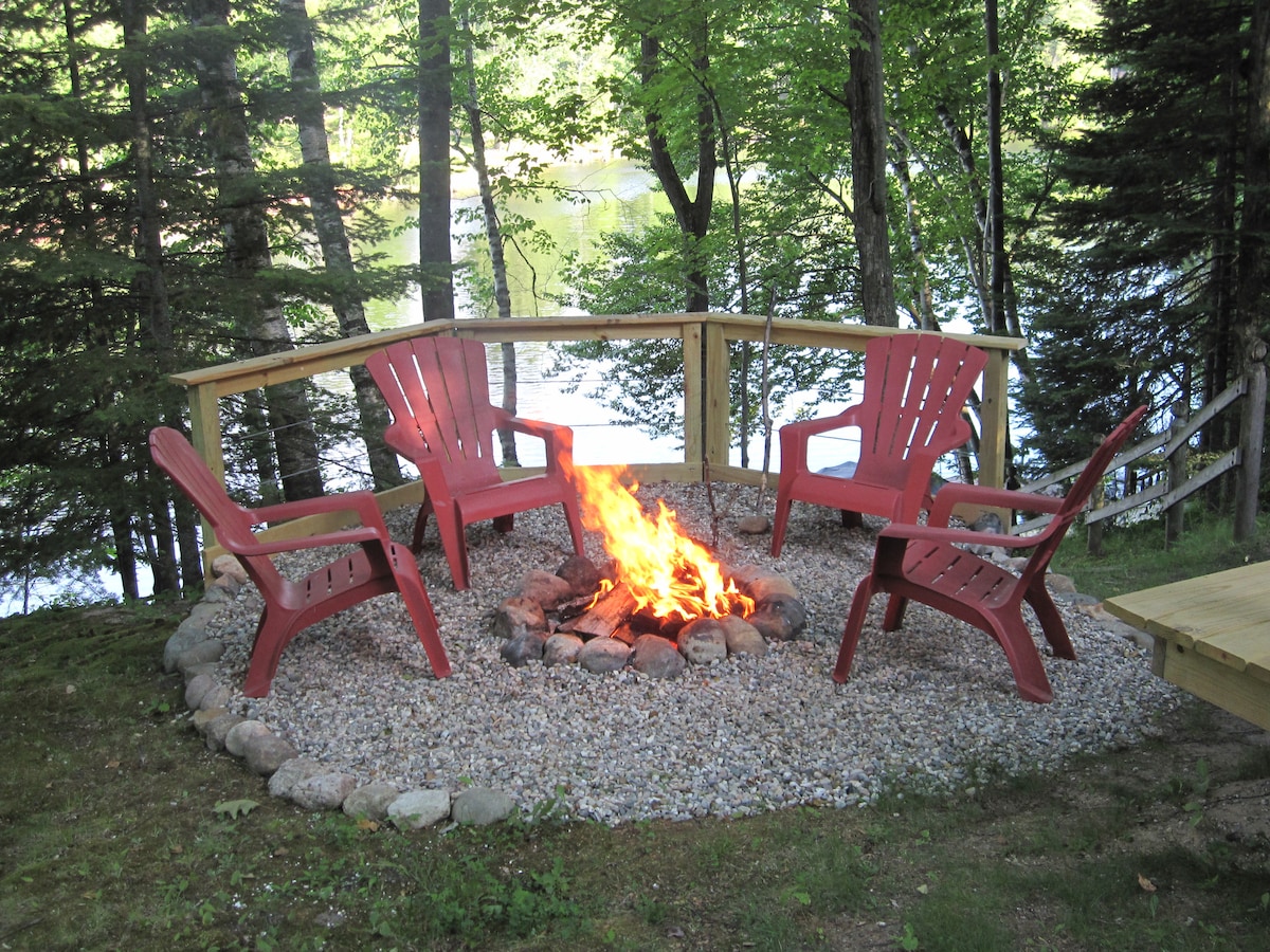 A fire pit surrounded by four red Adirondack chairs is situated on a gravel base, nestled within a forested area. A wooden railing provides boundaries, while trees offer natural shade. Flickering flames rise from the center of the pit, with a glimpse of the lake visible in the background.