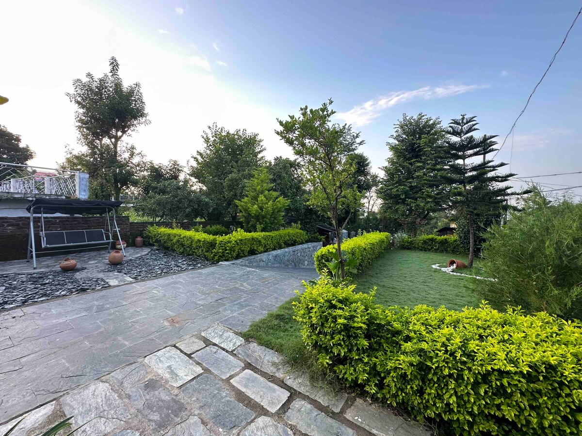 A landscaped outdoor area is seen, featuring a lush green lawn bordered by neatly trimmed hedges. Pathways of stone lead through the garden, which is dotted with trees and decorative plants. A seating area is visible under a shade structure, inviting relaxation.