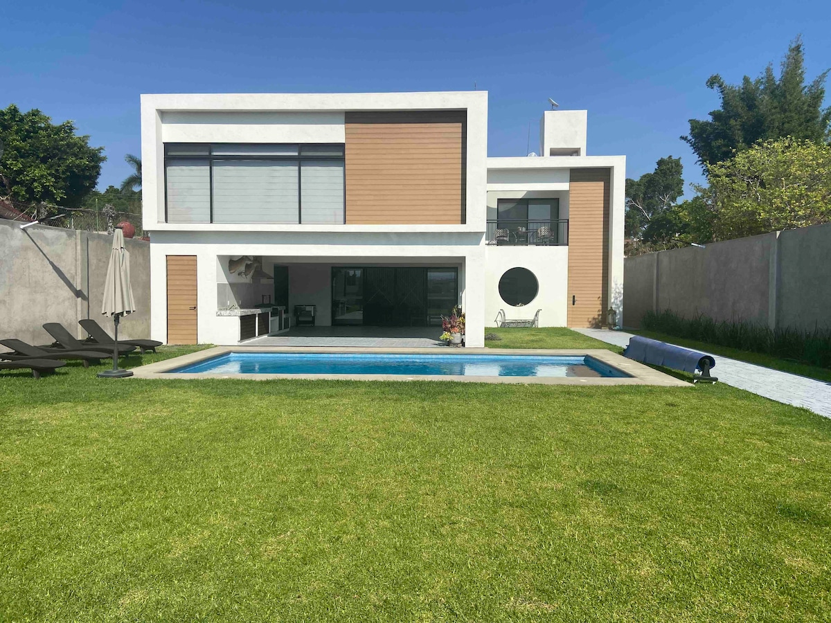 A modern two-story house is presented, featuring a spacious lawn and a private pool. Large windows enhance the facade, while a circular window adds architectural interest. The pool layout is designed for leisure, surrounded by lounge chairs, and lush greenery frames the property.