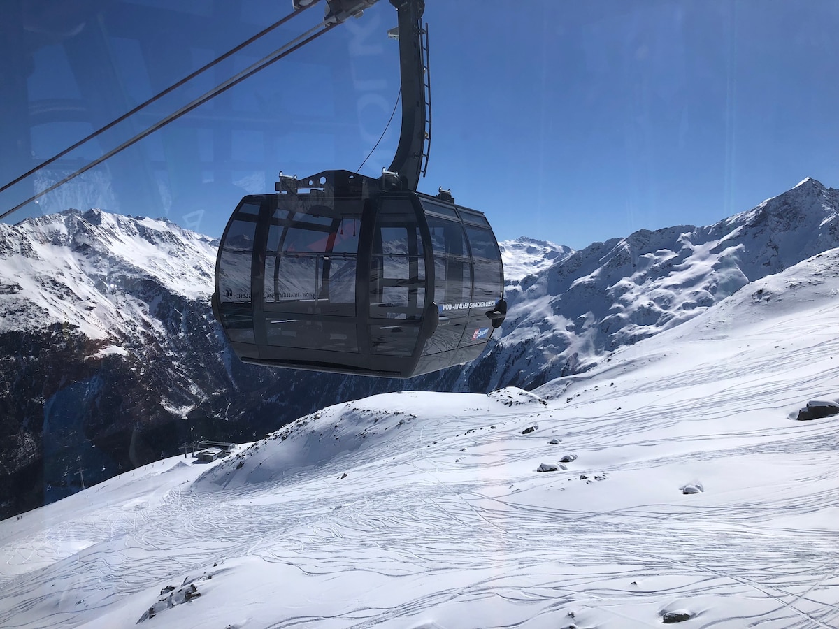 A gondola is shown ascending through a snowy landscape, surrounded by majestic mountains. The bright blue sky contrasts with the snow-covered terrain, highlighting the vastness of the area. Tracks left by skiers are visible on the pristine snow, adding to the serene atmosphere.