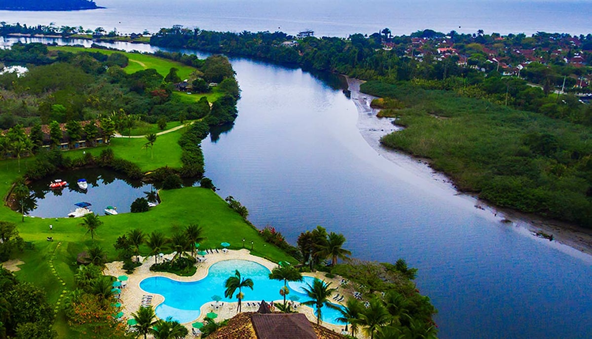 An aerial view captures the expansive green landscape bordered by a winding river and a serene pool area. Lush palm trees surround the pool, while the distant shoreline features a blend of homes and natural vegetation, set against the backdrop of the ocean.