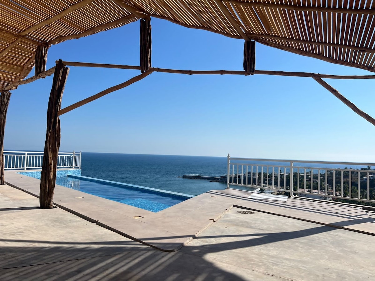 A spacious terrace features a stunning ocean view, with a shimmering infinity pool visible nearby. The serene blue sky meets the water's horizon, while sunlight casts gentle shadows across the floor. Natural wood beams provide a structural element, enhancing the outdoor space.