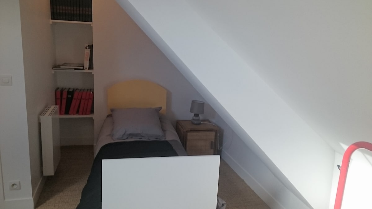A compact sleeping area showcases a single bed with a light-yellow headboard, positioned against a slanted wall. A bedside table is accompanied by a small lamp. Bookshelves filled with red-covered books are visible in the corner, adding a touch of color to the room.