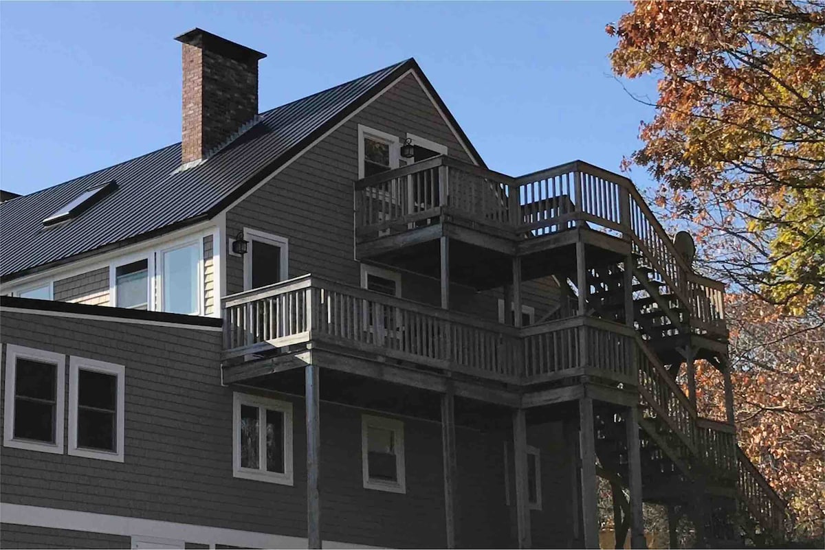 The exterior of a two-story building is displayed, featuring a gray facade with white trim. A wooden deck wraps around the second floor, providing multiple seating options. Stairs lead to the upper deck, where a satellite dish is visible. The clear blue sky complements the autumn foliage.