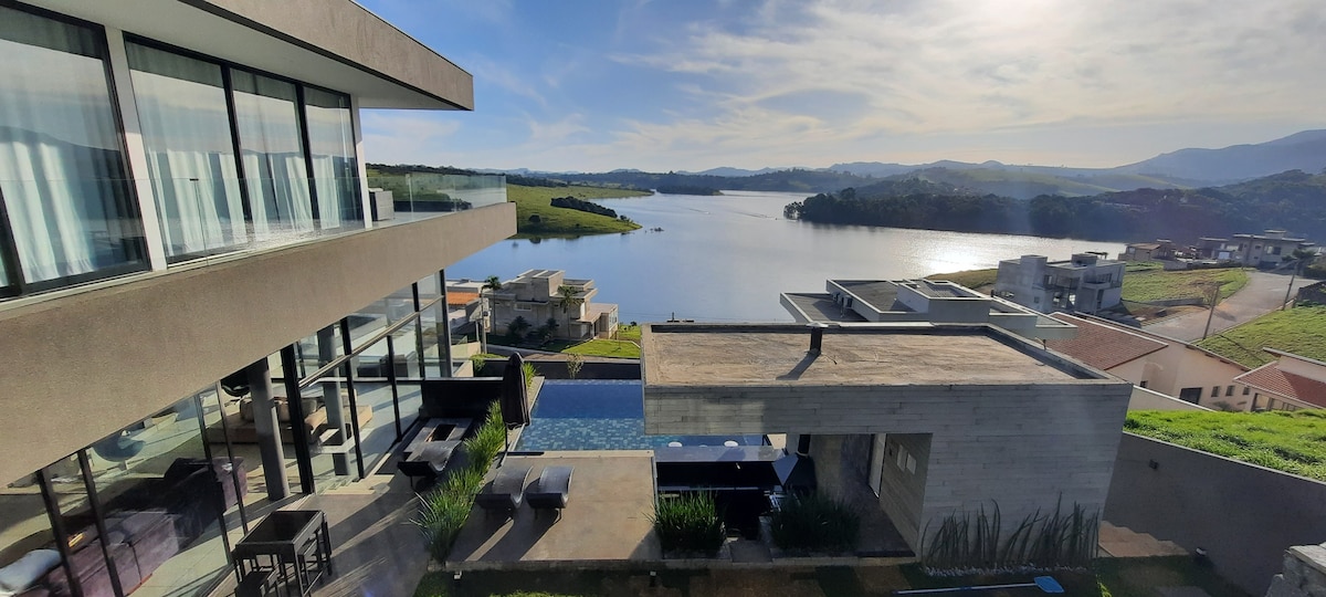 The contemporary home features large glass walls, offering panoramic views of the tranquil Jaguari reservoir. The infinity pool extends toward the water, blending seamlessly with the landscape. Lush greenery and modern architecture enhance the serene outdoor space, inviting relaxation and enjoyment of the picturesque surroundings.