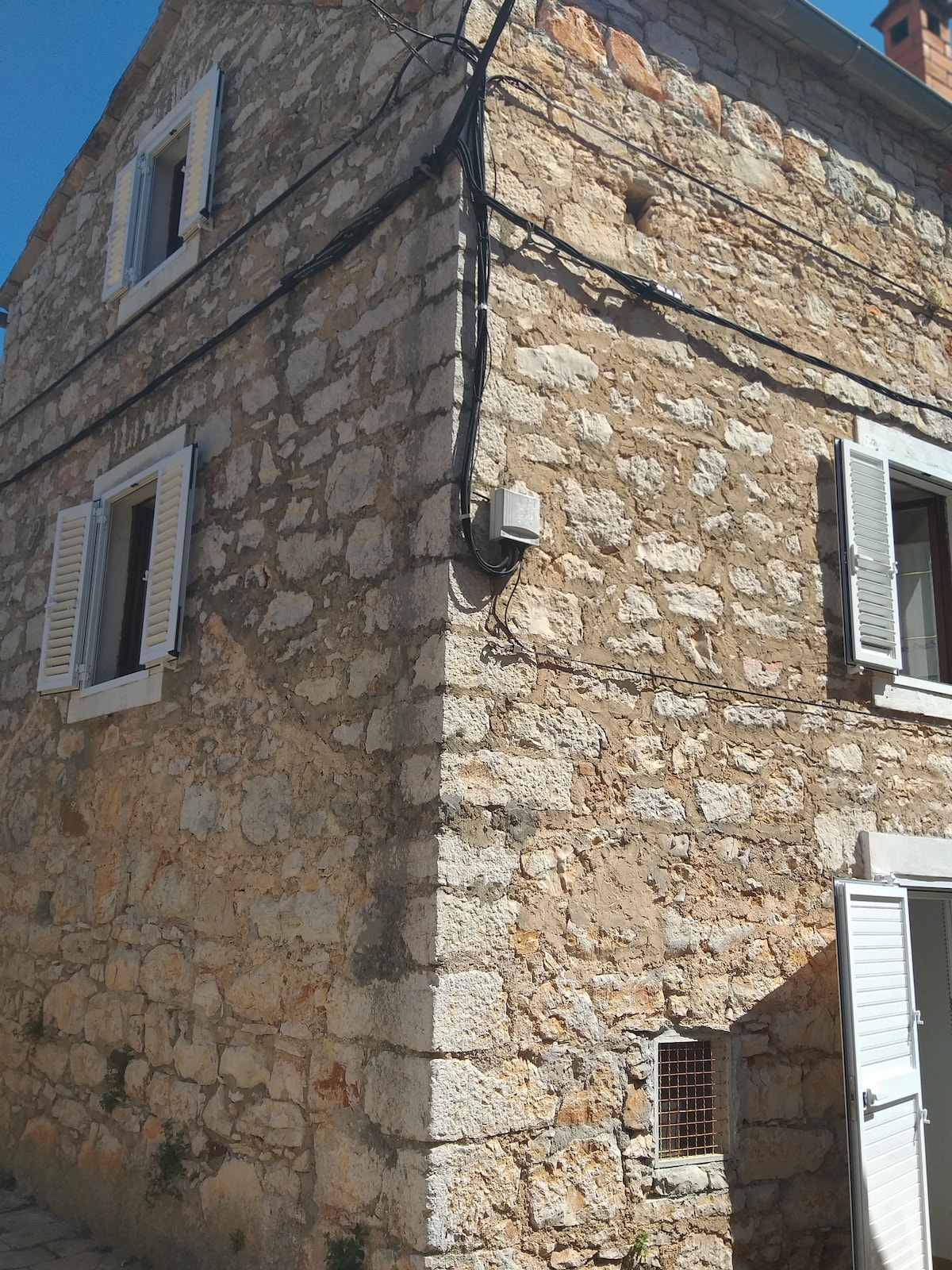 The exterior of a stone house features a rustic texture with a combination of warm earth tones. White shutters adorn the windows, while an accessible entrance and an electrical unit are positioned at the side. The image captures a tranquil street environment.