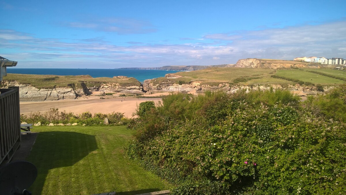 A panoramic view of Porth Beach and the north coast of Cornwall is presented, showcasing a blend of sandy shoreline, rocky cliffs, and lush green landscapes. The sea reflects clear blue skies, while distant buildings can be seen along the coastline, contributing to the natural setting.