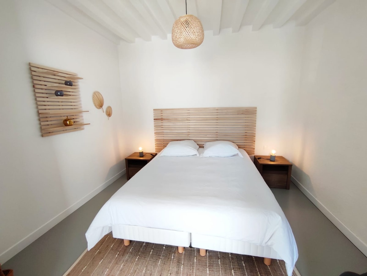 A bright bedroom features a spacious bed with a light, neutral-colored duvet. Wooden nightstands are positioned on either side, each illuminated by a small lamp. Minimalist wall decorations, including woven elements, enhance the serene atmosphere.