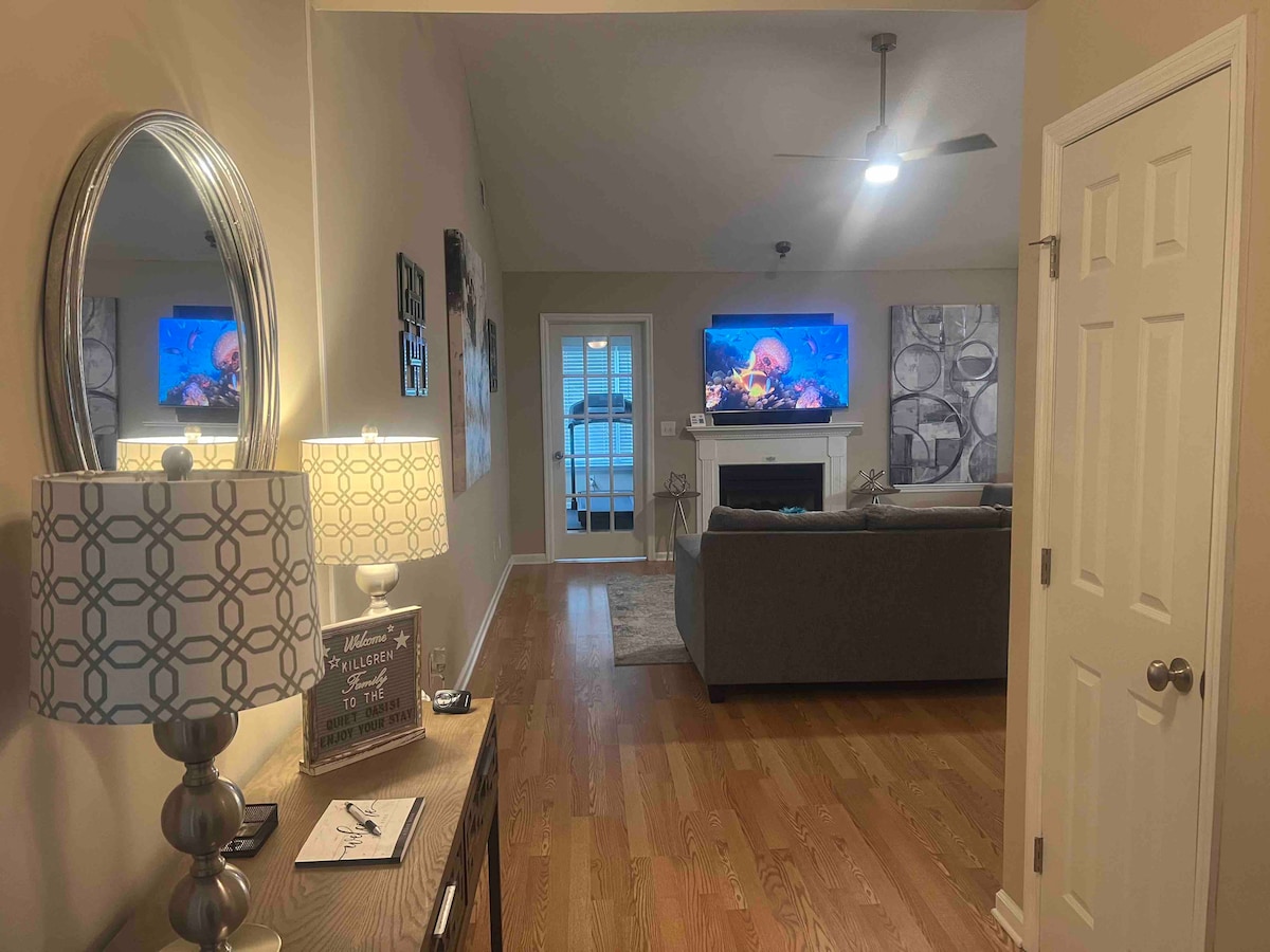 A welcoming entryway leads into a spacious living area, highlighted by a modern light fixture beside a decorative console table. Two framed artworks adorn the walls, while a large couch faces two mounted TVs above a fireplace. Natural light enters from a sliding glass door.