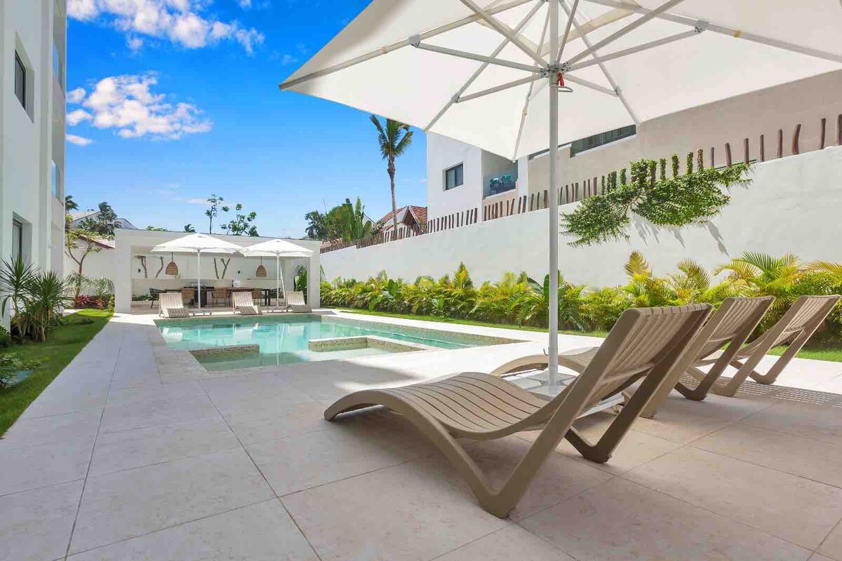 A serene pool area is featured, surrounded by lush greenery. Three lounge chairs are positioned under a large umbrella, offering shaded seating. White cabanas provide additional relaxation space near the sparkling water, all set against a clear blue sky.
