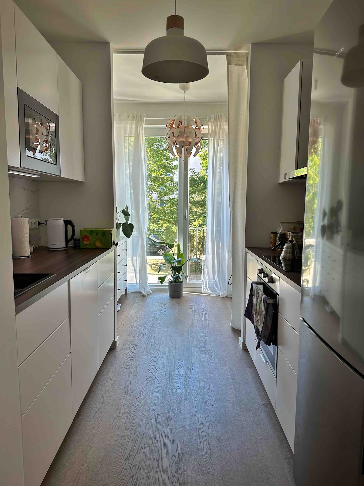 A clean, modern kitchen features sleek white cabinetry and a dark countertop. Large windows adorned with sheer curtains allow natural light to brighten the space. A potted plant is positioned near the window, enhancing the fresh and inviting atmosphere.