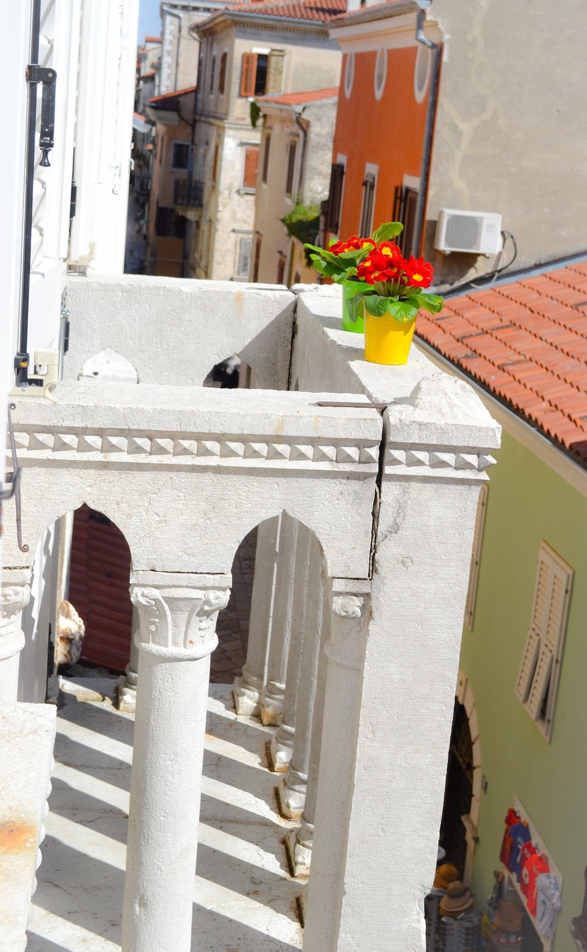 A balcony view showcases charming old town buildings with colorful facades and tiled roofs. A yellow planter filled with vibrant flowers adds a pop of color to the stone railing. The sunlight casts gentle shadows across the architectural details of the balcony.