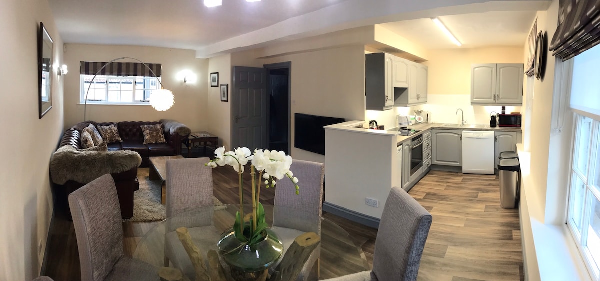 An open-plan kitchen and dining area features a modern design with a circular dining table at the forefront. A cozy sofa is positioned nearby, complemented by a stylish floor lamp. The well-equipped kitchen is visible in the background, with light-colored cabinets and appliances integrated into the space.