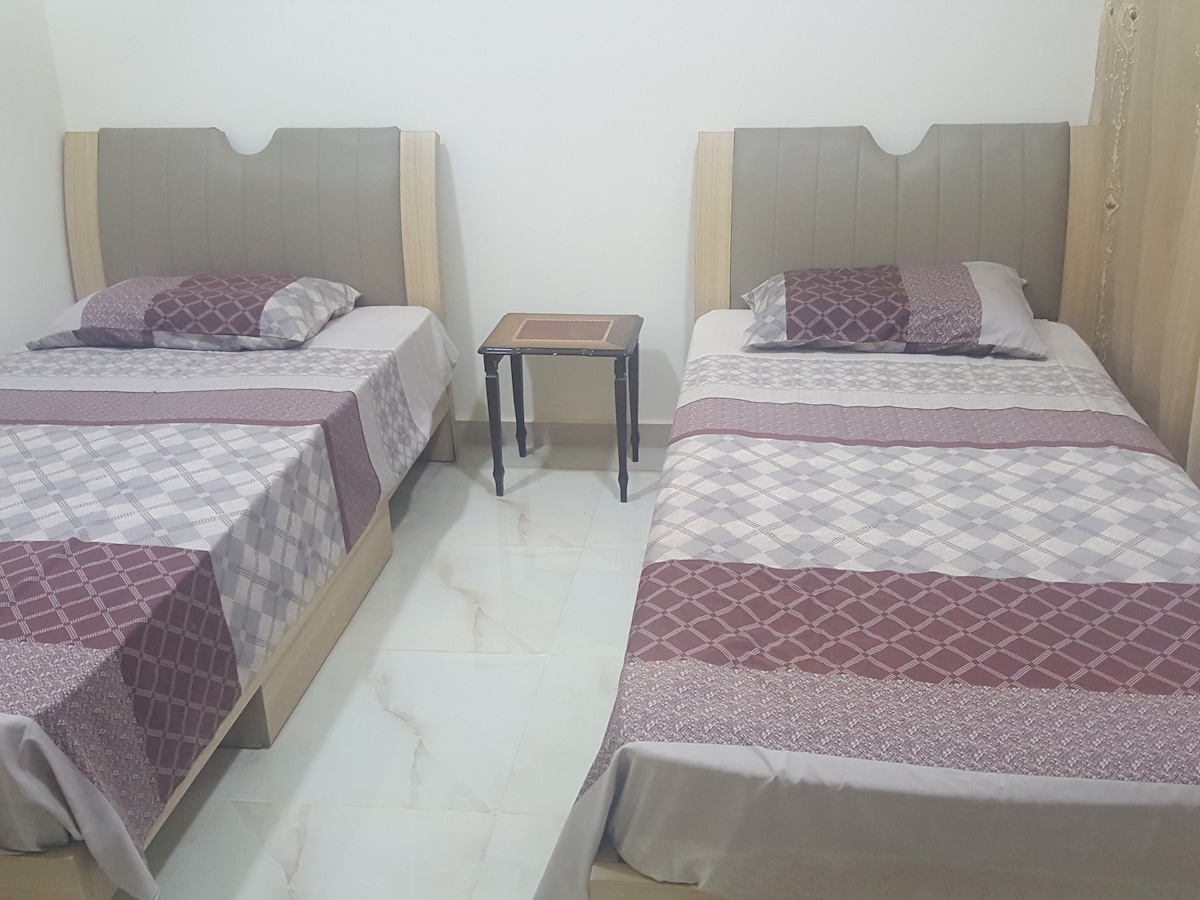 A clean sleeping area features two spacious beds placed parallel to each other, each with patterned bed coverings in muted tones. A small, dark wood table stands between the beds, adding practicality to the room's layout.