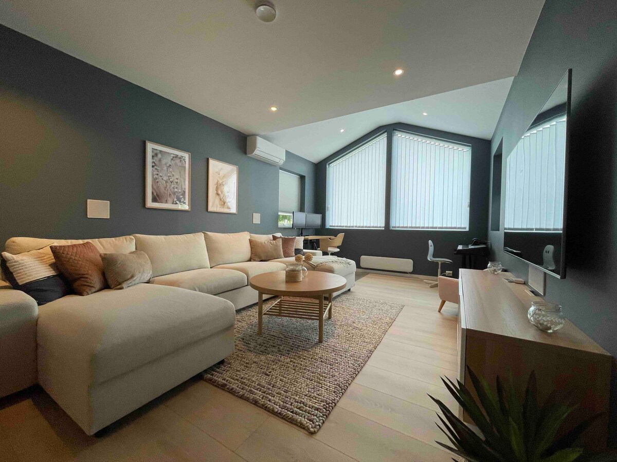 A modern living area features a spacious sectional sofa complemented by decorative cushions. A round coffee table sits on a textured rug, while natural light filters through large windows with vertical blinds. A small workspace with a desk and chair is visible in the background.