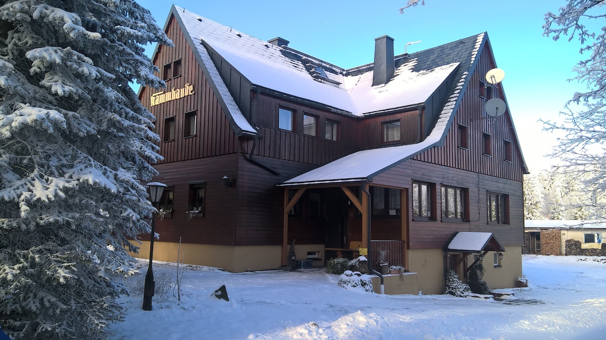 A large wooden building is set against a snowy landscape, featuring multiple windows and a covered porch. The entrance is highlighted, with snow-covered trees surrounding the property. Satellite dishes are visible on the roof, emphasizing the rustic charm of the structure.