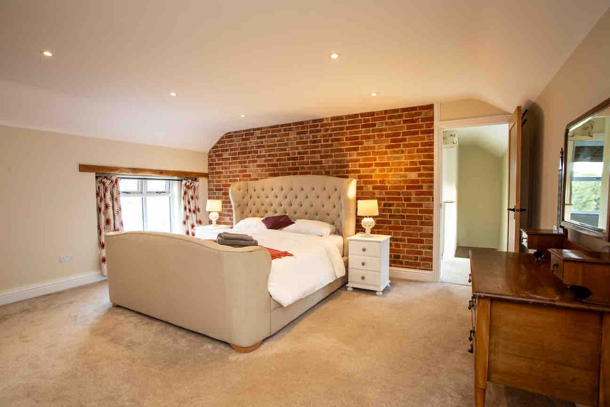 A spacious bedroom features a plush bed with a tufted headboard, flanked by bedside tables with lamps. A rustic brick wall adds character, while windows with patterned curtains provide natural light. A wooden dresser complements the warm tones of the room.