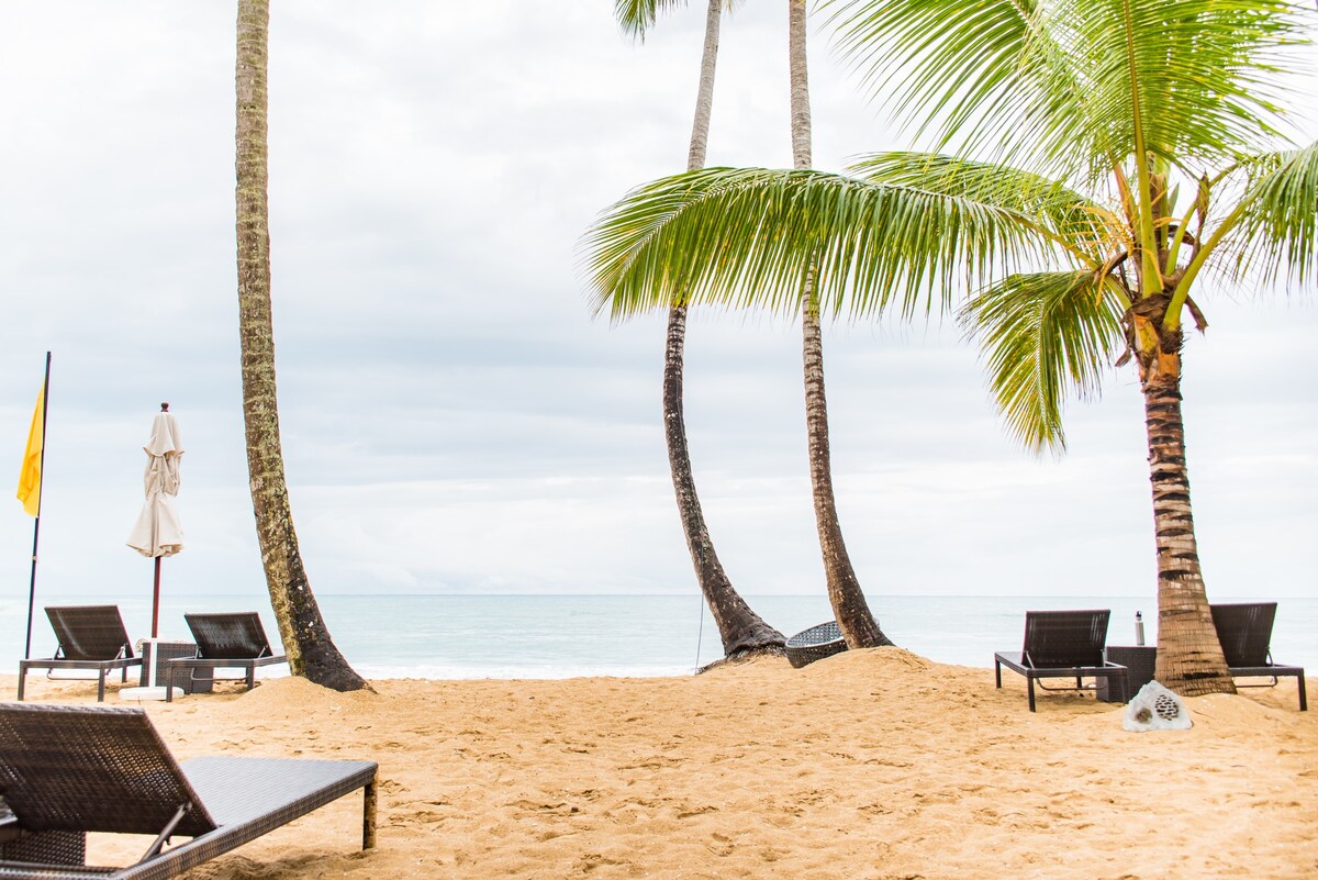 A sandy beach area is framed by tall palm trees, offering a relaxed atmosphere. Several lounge chairs are arranged in the sand, facing the calm ocean. A beach umbrella is positioned nearby, providing shaded space for relaxation.