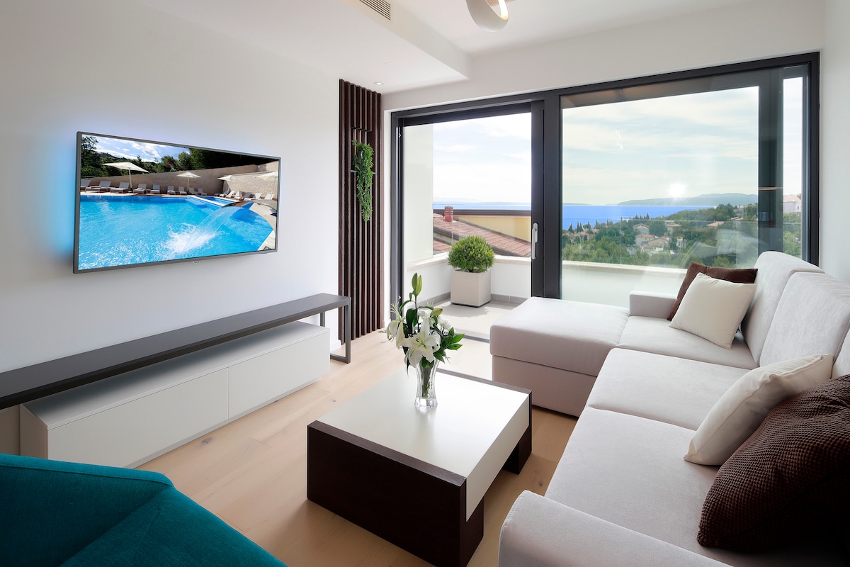 A modern living area features a light-colored sofa with a neutral palette and a coffee table. A flat-screen TV is mounted on the wall, showing a clear view of an outdoor pool area. Large glass doors offer expansive views of the outdoor landscape.