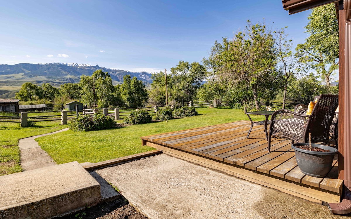 A wooden deck offers a serene outdoor space, featuring comfortable seating positioned to overlook sprawling green fields and distant mountains. Lush trees and well-maintained landscaping can be seen in the surrounding area, creating a peaceful rural setting.