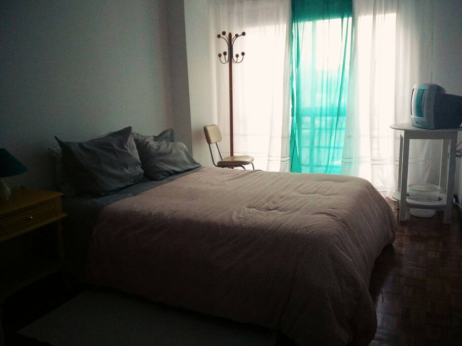 Quarto acolhedor com cabide de chão, mesa de cabeceira e candeeiro, cadeira, televisão, roupeiro e espelho