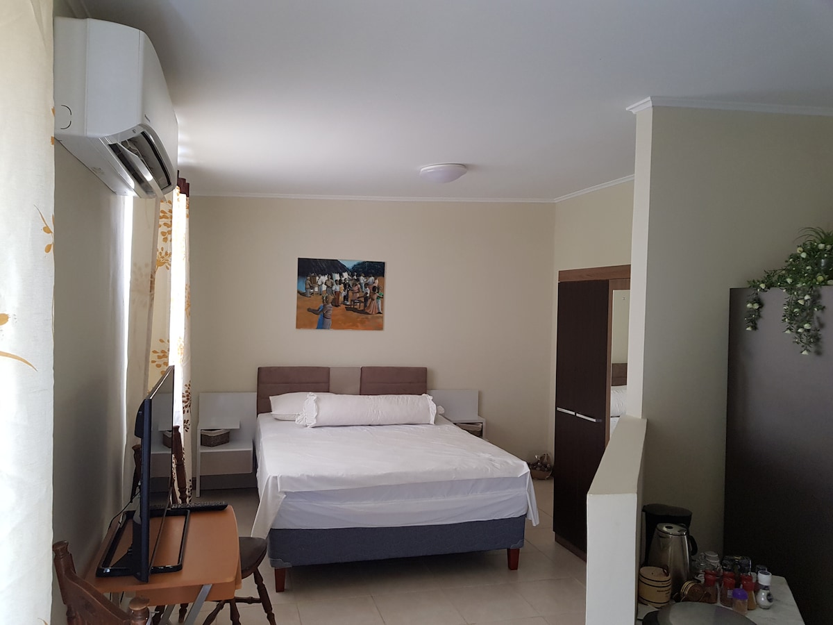 A spacious bedroom features a large bed with neutral bedding. A television is positioned on a wooden table nearby. Light enters through large curtains, brightening the room. An artwork depicting people in a social setting adorns the wall above the bed.