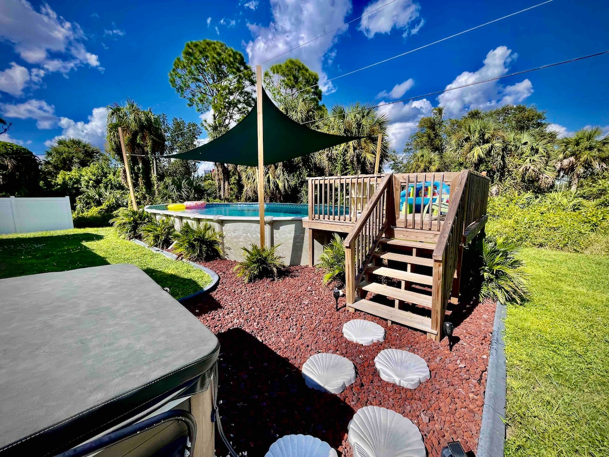 The outdoor space features a wooden deck leading to a heated above-ground pool, shaded by a large canopy. Lush greenery surrounds the area, with decorative stones and four white seating shells arranged on the ground, creating a relaxing atmosphere.