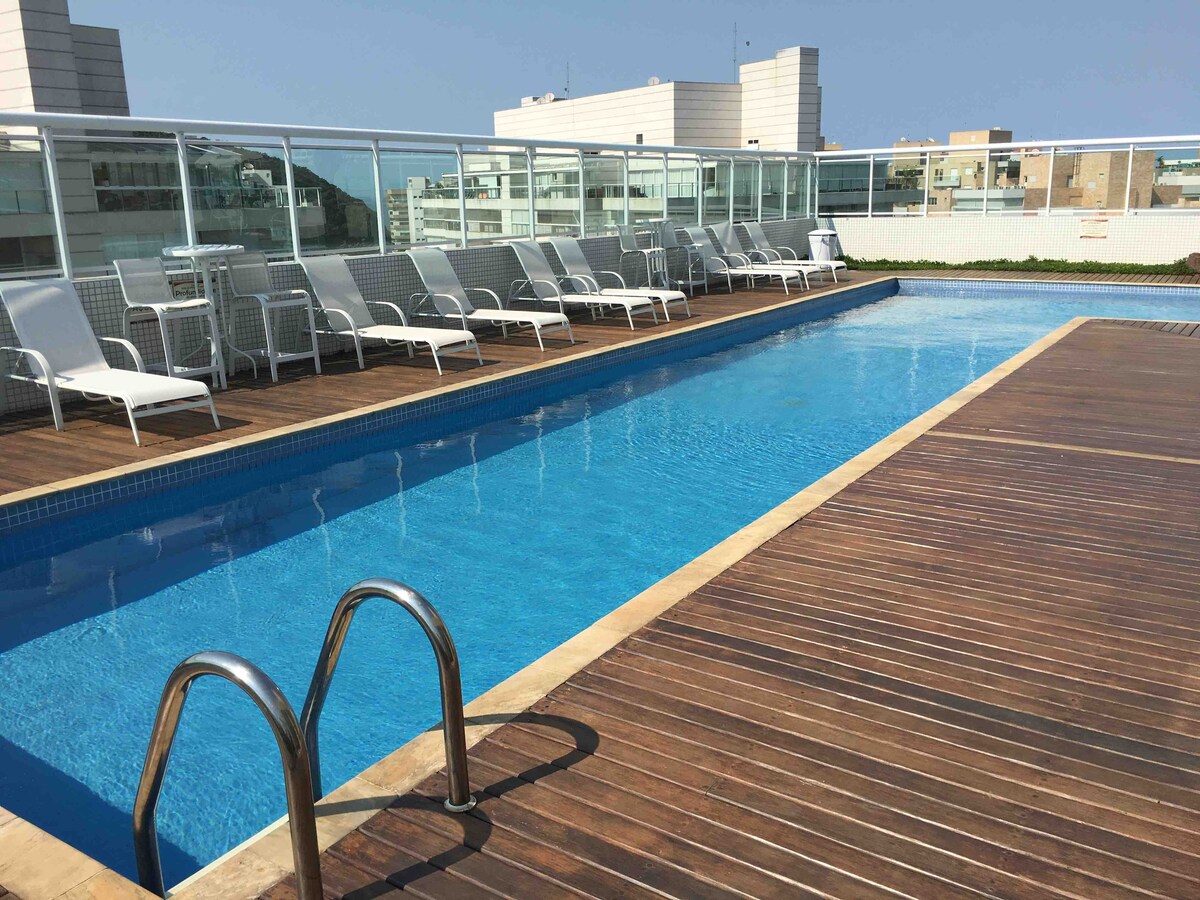 A rooftop pool area features a sleek, rectangular swimming pool surrounded by a wooden deck. There are several lounge chairs arranged along the edge, offering a place to relax. The backdrop includes distant buildings under a clear blue sky.