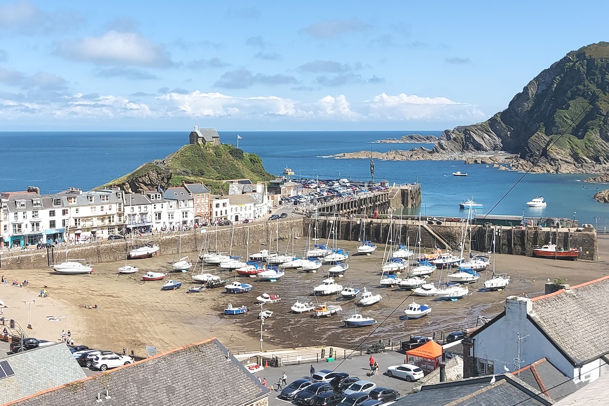 A panoramic view of Ilfracombe's working harbour is depicted, showcasing several boats moored in the water and the surrounding scenic coastline. The vibrant blue sea contrasts with the sandy beach, while buildings line the waterfront, providing a sense of community near the bustling port.