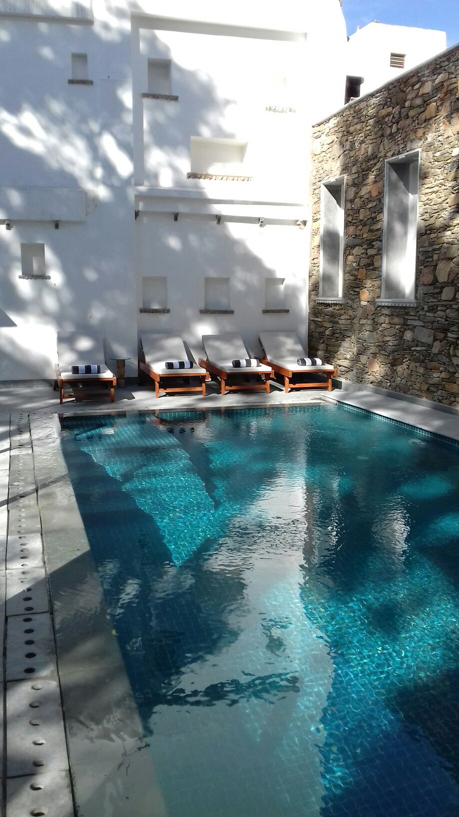 A serene swimming pool is surrounded by smooth stone walls, reflecting gentle sunlight. Several lounge chairs are positioned along the edge, inviting relaxation. The pool's turquoise water glimmers, showcasing a tranquil atmosphere within the shared courtyard setting.