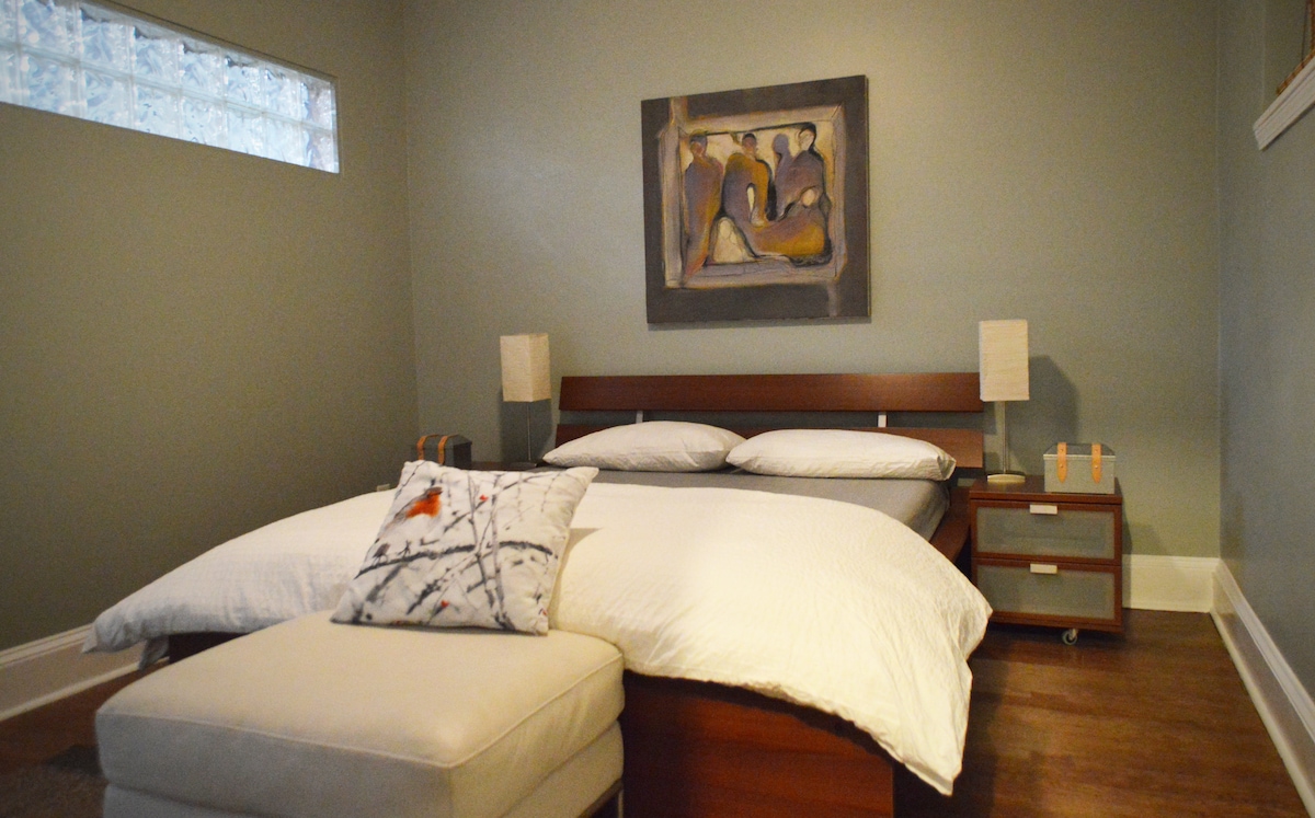 The bedroom features a spacious bed with neatly arranged bedding, accompanied by two bedside lamps on wooden nightstands. A plush ottoman is positioned at the foot of the bed. A distinctive artwork hangs above the headboard, while natural light filters through a textured glass block window.