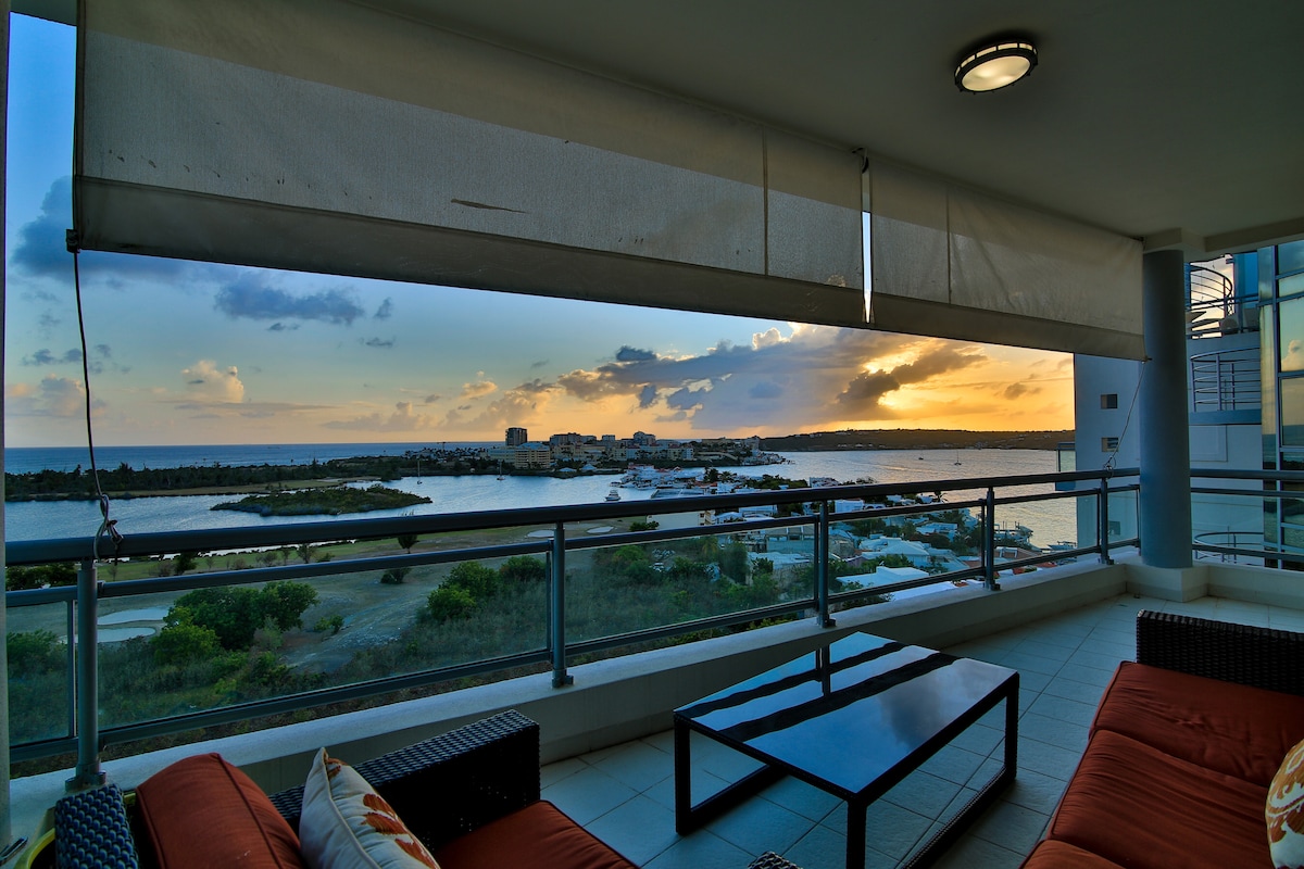An outdoor seating area features a modern design with comfortable sofas and a coffee table. Panoramic views of the lagoon and the Caribbean Sea are seen at sunset, with clouds illuminated in warm tones. Large windows offer an unobstructed view of the serene landscape.