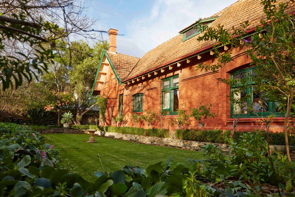 The exterior of the historic apartment is showcased, featuring a warm-toned brick facade with decorative green window frames. Lush green grass and well-maintained garden beds filled with plants surround the building, creating a tranquil outdoor setting.